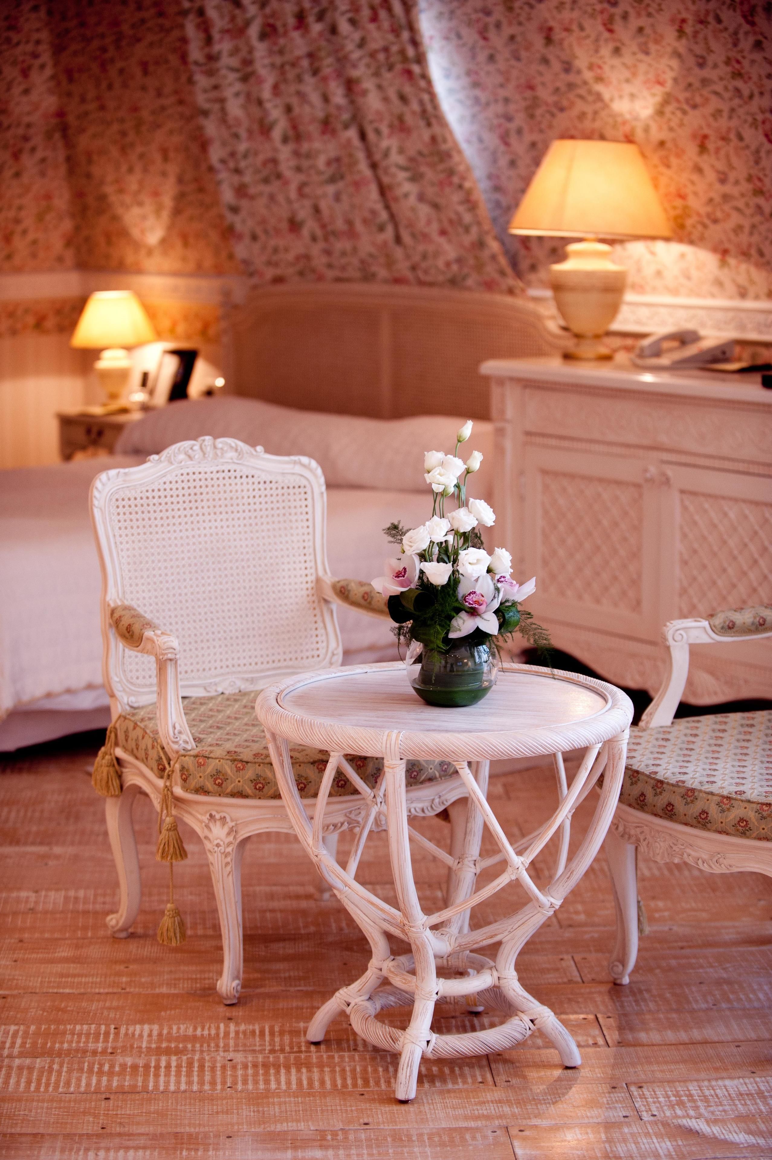 A classic room with a bed, two chairs, a round table with a vase of flowers, and two lamps on nightstands.