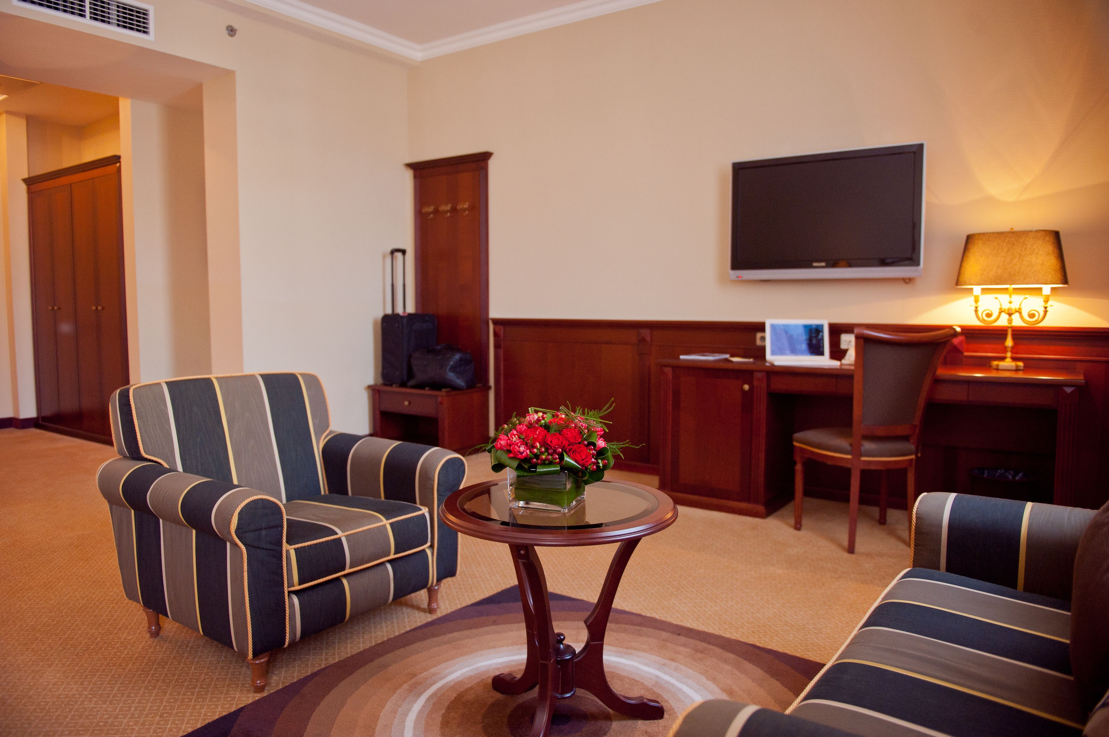 A hotel room with striped armchairs, a small round table with a vase of flowers, a desk with a computer, and a flat-screen TV mounted on the wall.