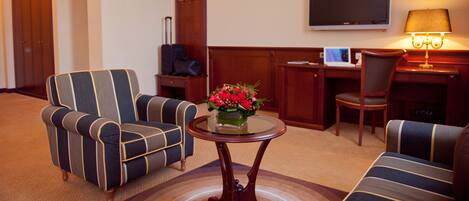 A hotel room with striped armchairs, a small round table with a vase of flowers, a desk with a computer, and a flat-screen TV mounted on the wall.
