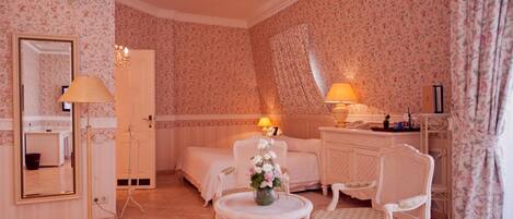 A room with floral wallpaper, a bed, a bedside table with a lamp, a chair, and a small table with a vase of flowers.