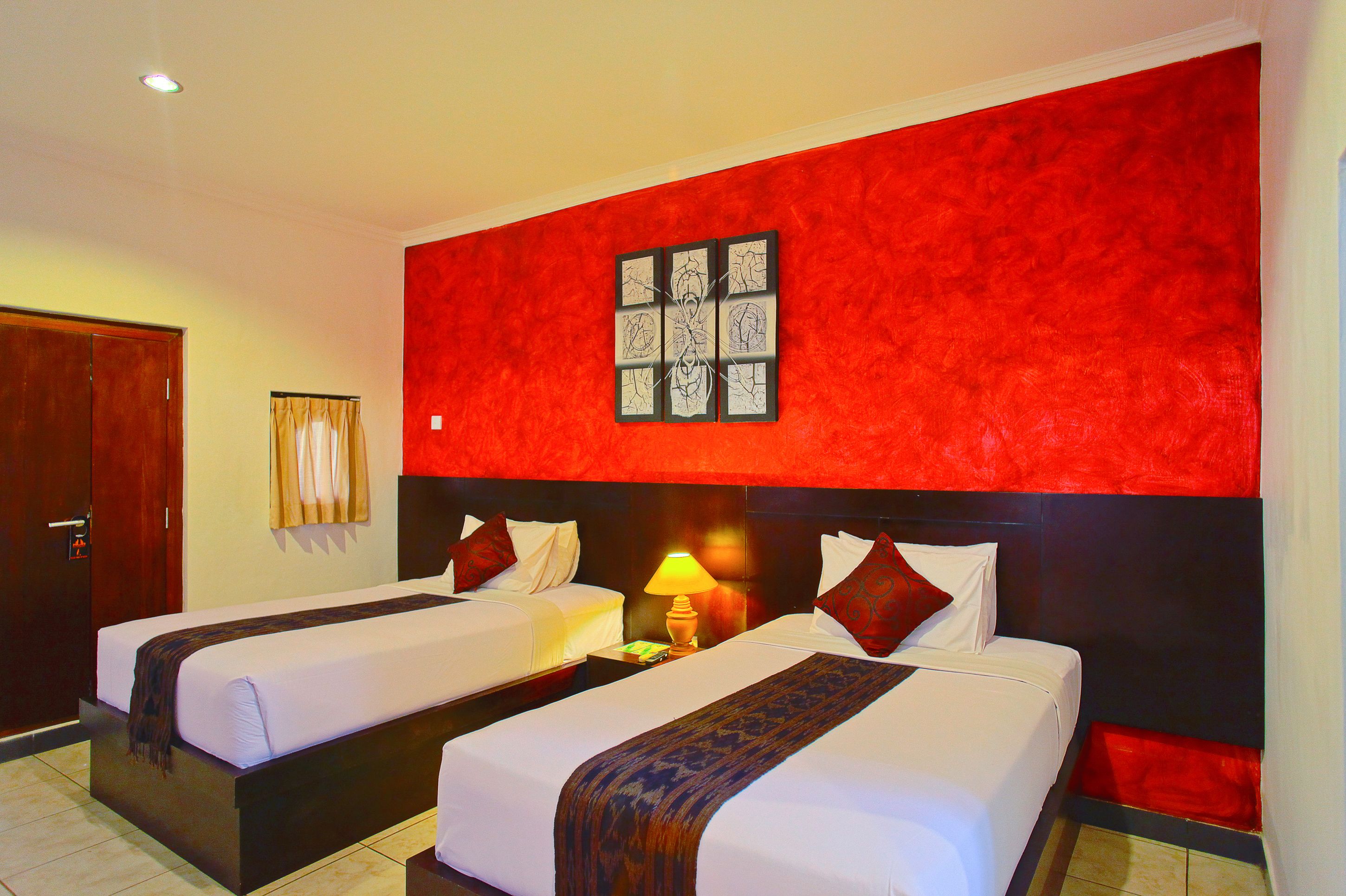 A hotel room with two beds, a red accent wall, a framed artwork, and a nightstand with a lamp.