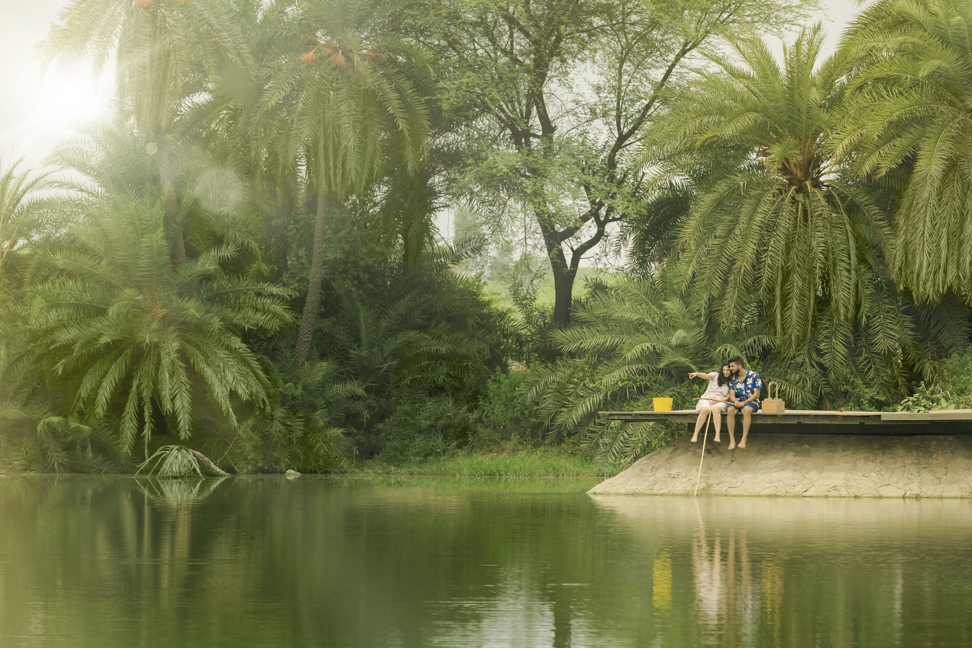 Un couple est assis sur une bordure en béton, au bord d’une étendue d’eau calme, entouré d’une végétation luxuriante et de palmiers.