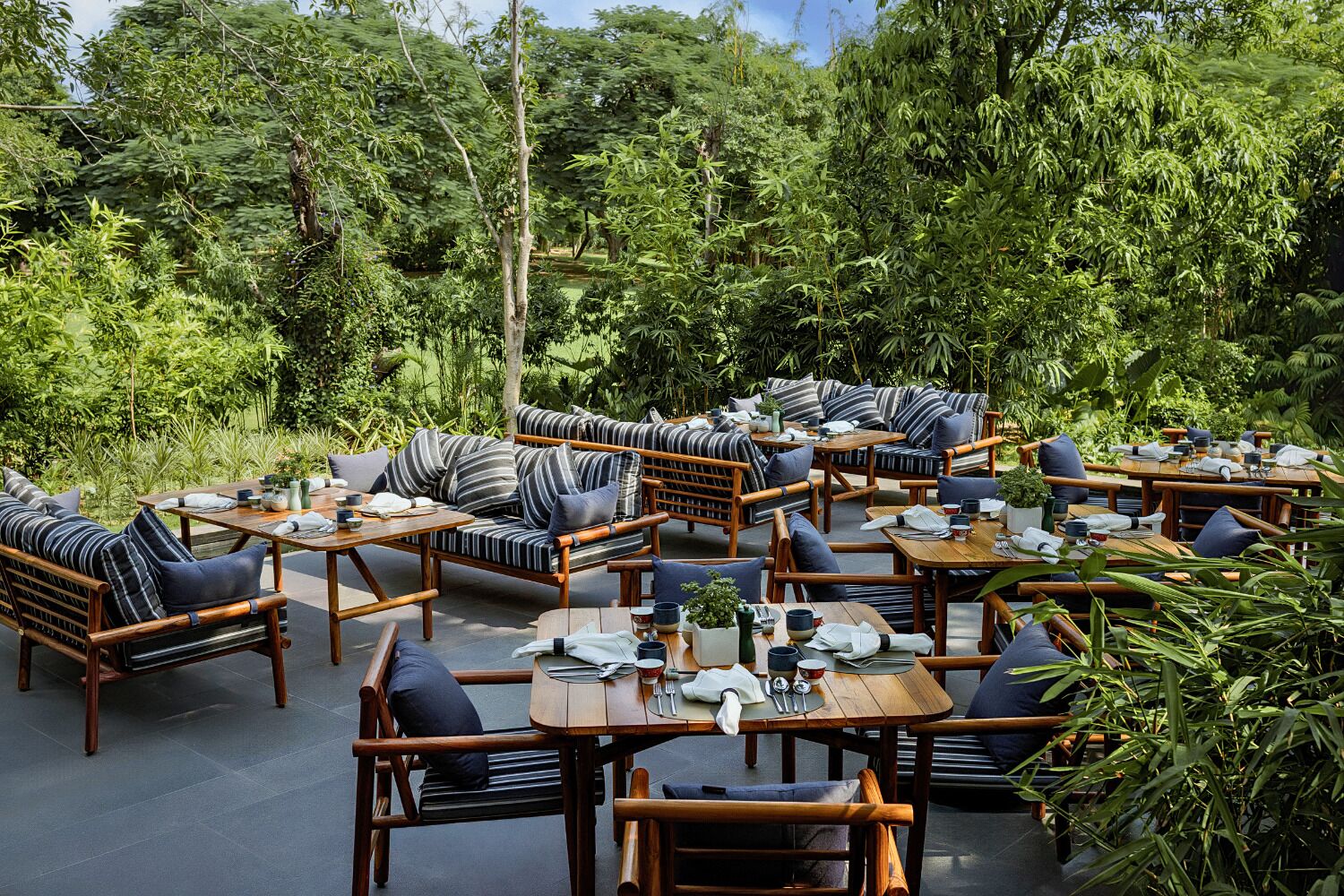 An outdoor seating area with wooden furniture and cushions, surrounded by lush greenery.