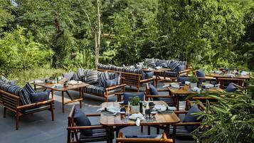An outdoor seating area with wooden furniture and cushions, surrounded by lush greenery.