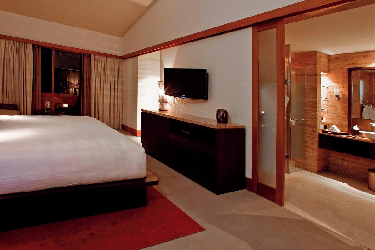 A hotel room with a large bed, a TV mounted on the wall, a desk with a lamp, and a bathroom with a mirror and sink.