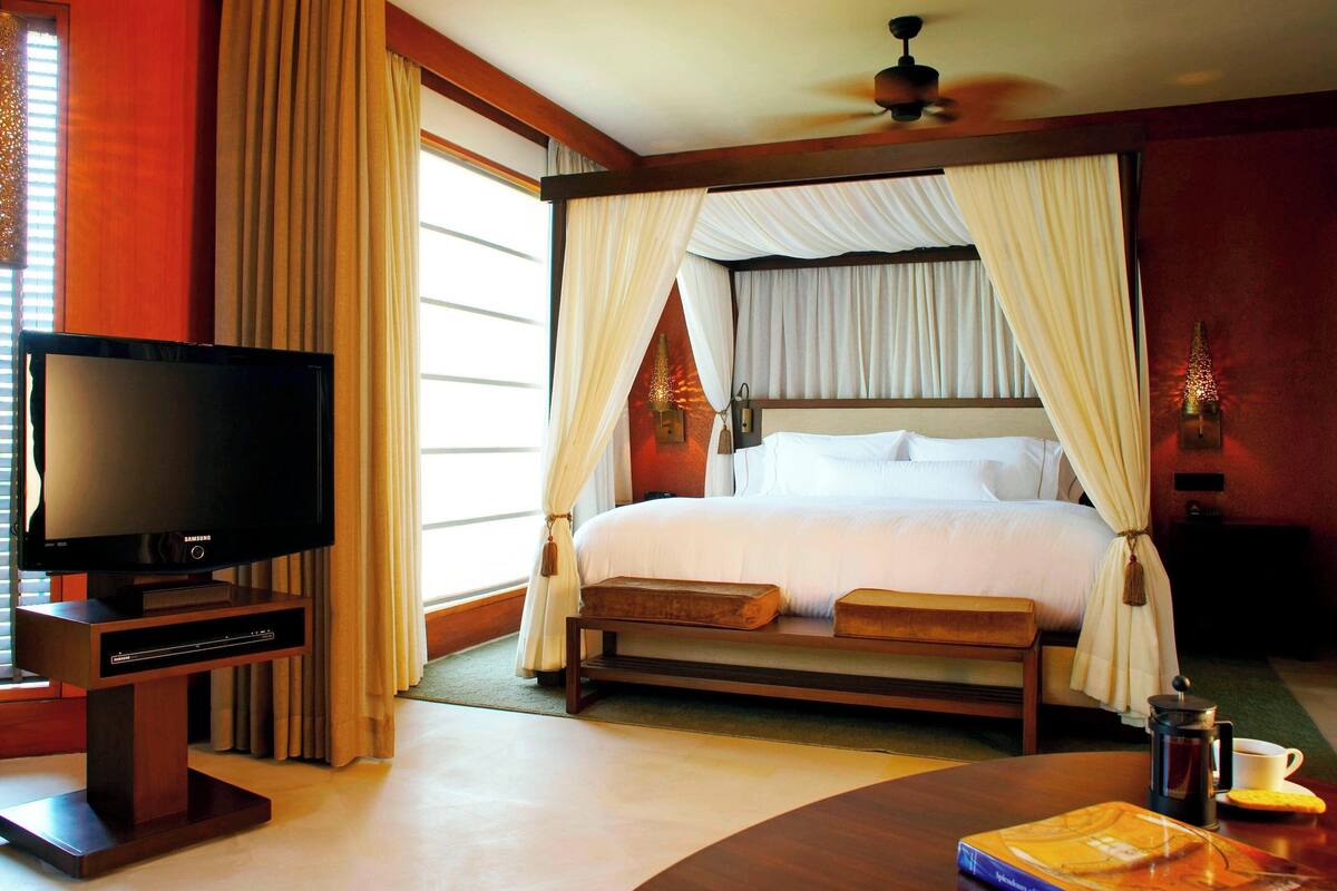 A bedroom with a four-poster bed, a television on a wooden stand, and a bedside table with a cup and a book.