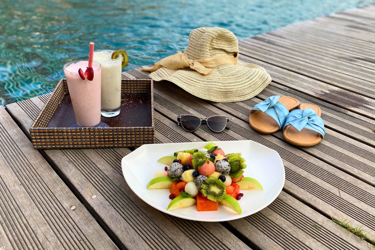 A wooden deck with a fruit platter, a drink tray, a straw hat, sunglasses, and flip-flops.