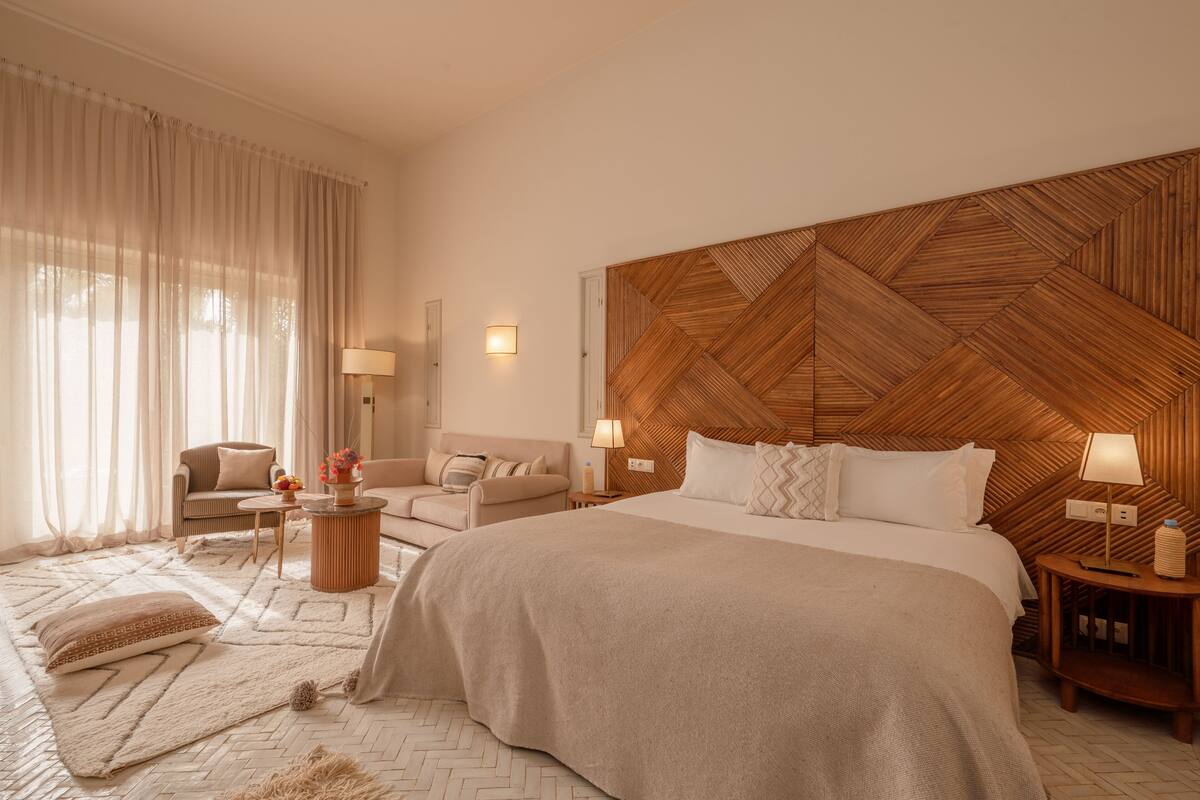 A spacious bedroom with a large bed, a wooden headboard, a sitting area with a sofa and armchair, and a dining table.