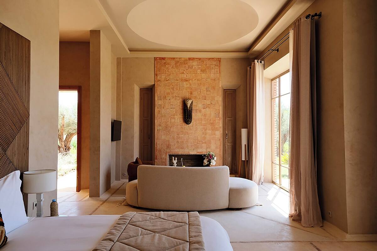 A spacious bedroom with a large bed, a curved seating area, a fireplace, and a view of the outdoors.