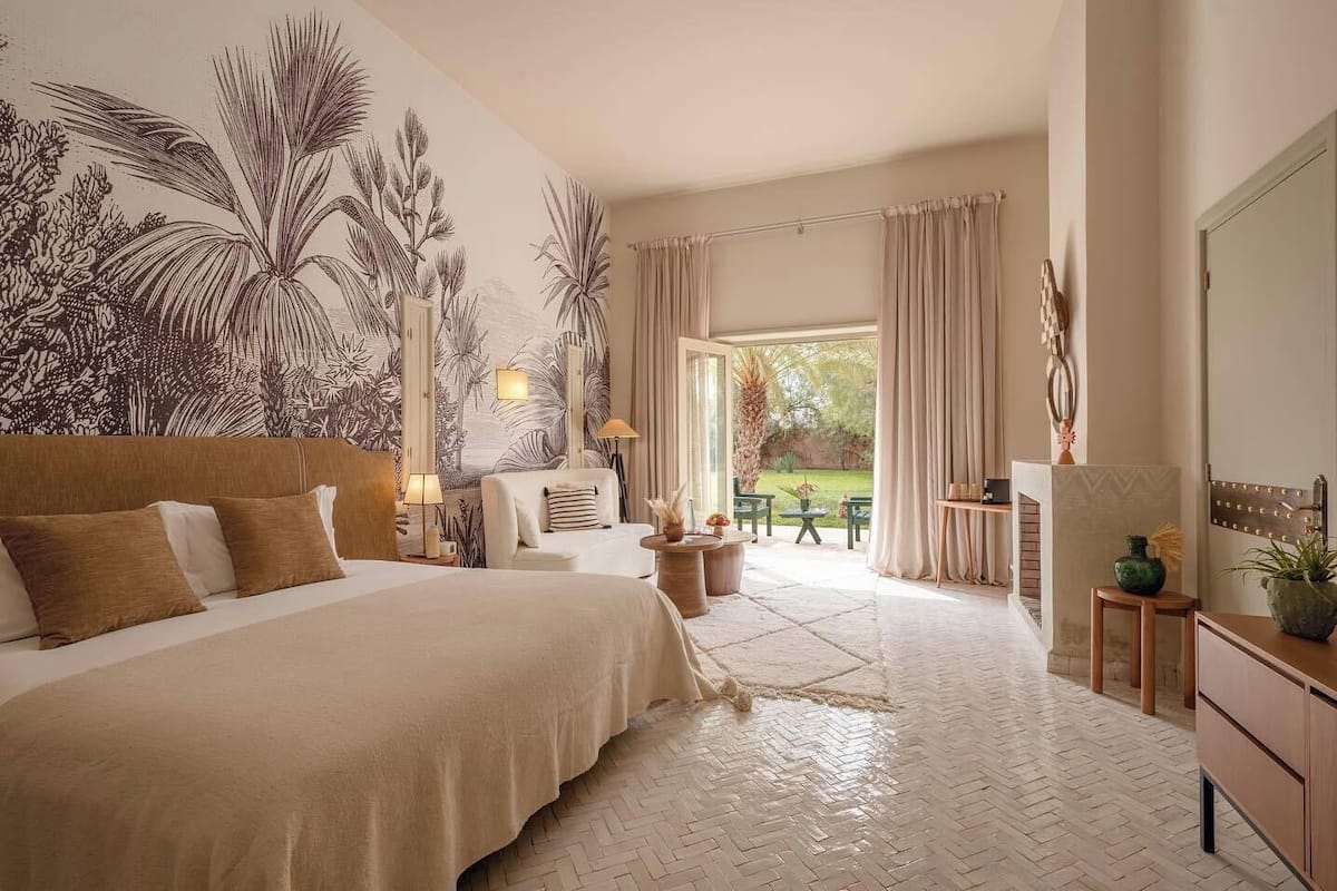 A spacious bedroom with a large bed, a seating area, a view of the garden, and a wall with tropical plant artwork.
