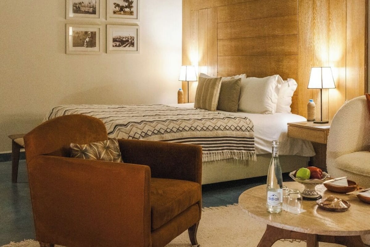 A hotel room with a bed, two bedside lamps, a brown armchair, a round coffee table, and a small table with a bottle and fruit.
