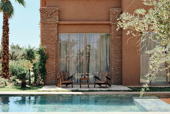 Zona junto a la piscina con cerramiento de vidrio, sillas de madera y una palmera.