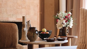 Una habitación con chimenea, una mesa redonda con un florero, un frutero y un jarrón rústico.