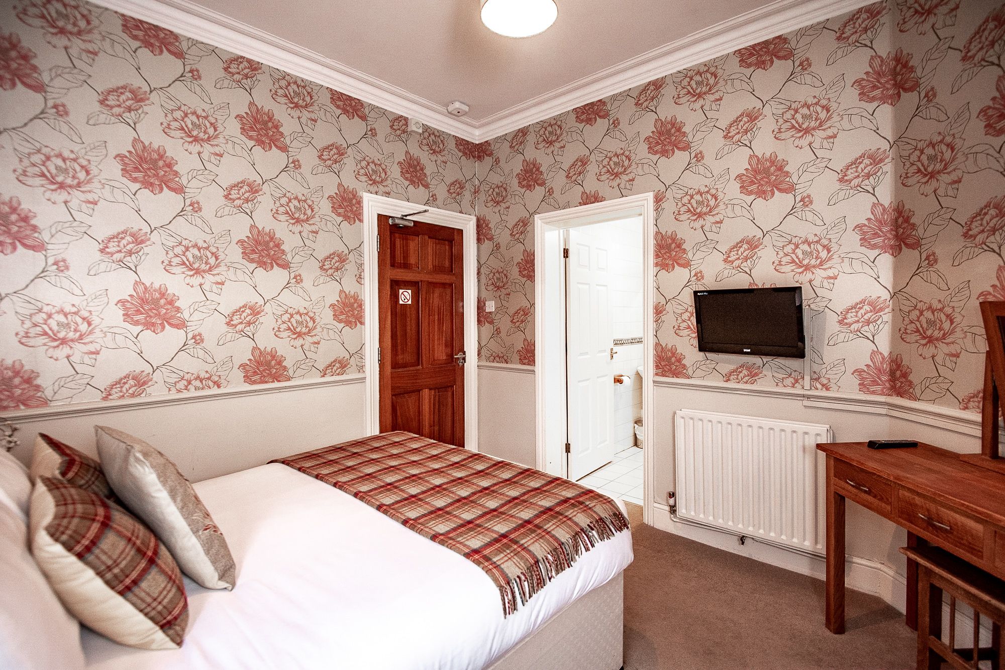 Un dormitorio con papel tapiz floral, una cama con manta escocesa, un aparador de madera y un televisor montado en la pared.