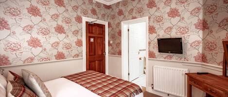 Un dormitorio con papel tapiz floral, una cama con manta escocesa, un aparador de madera y un televisor montado en la pared.