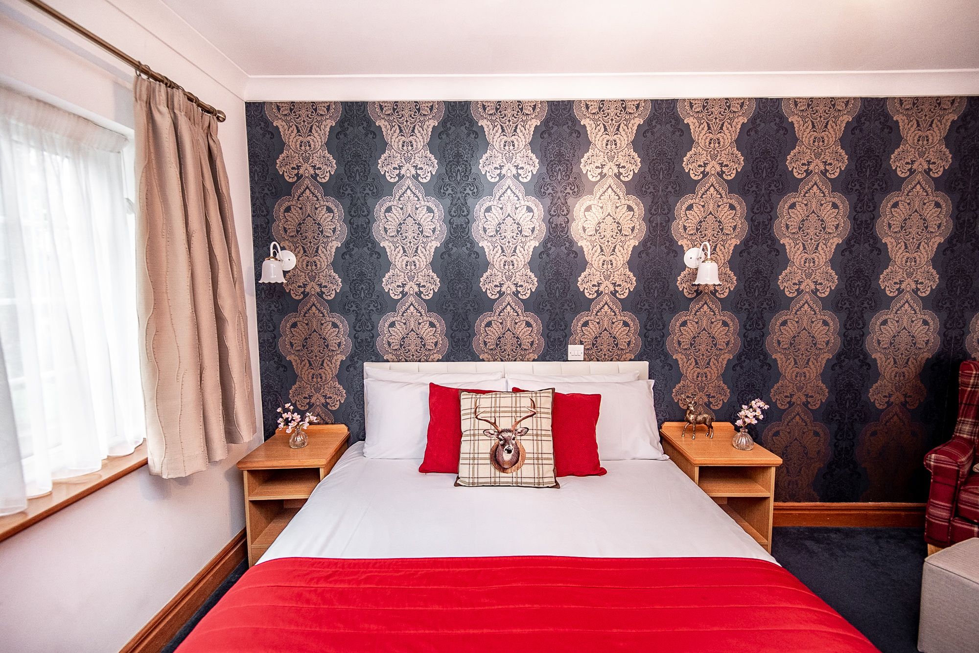 A hotel room with a large bed, two bedside tables, a nightstand, a red throw blanket, and a patterned wallpaper.