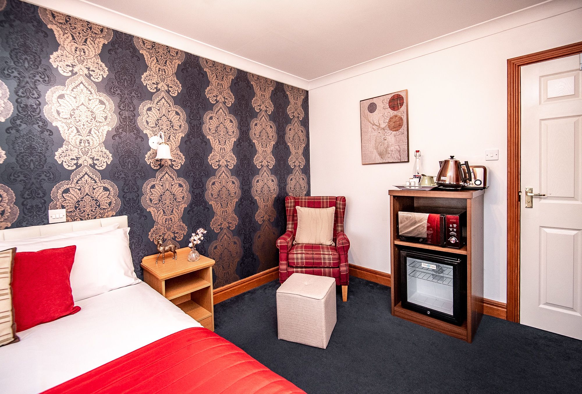 Une chambre d’hôtel avec un lit, une chaise rouge, un petit réfrigérateur et un mur recouvert d’un papier peint à motifs.