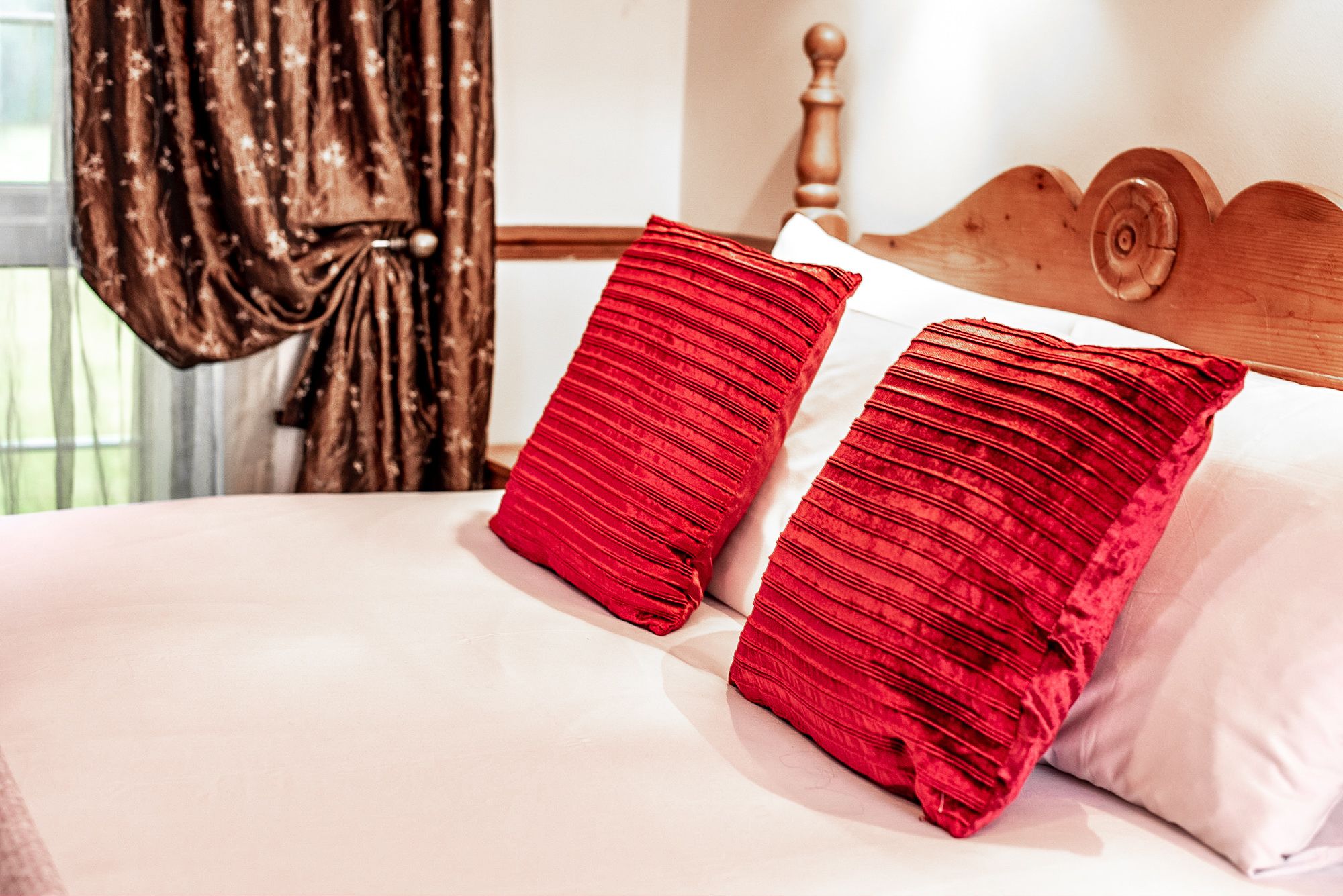 Una cama con dos almohadas rojas y una blanca, un cabecero de madera y una ventana con cortinas.