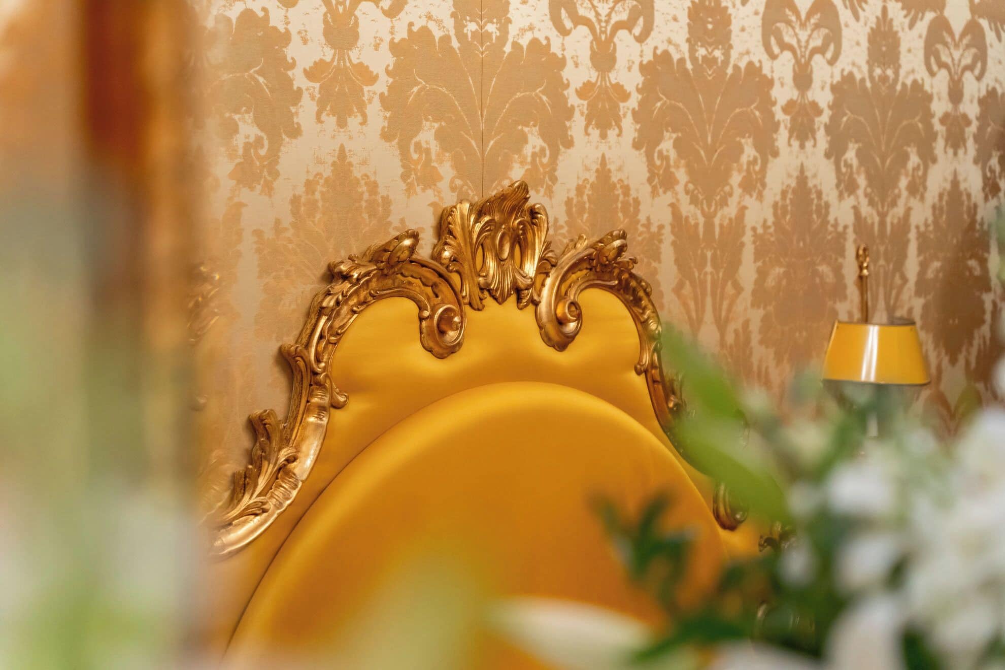 Une chambre avec une tête de lit dorée et décorée, un papier peint à motifs et une lampe jaune.