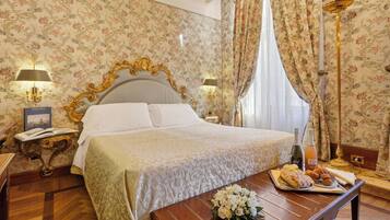 A hotel room with a large bed, floral wallpaper, a wooden floor, and a table with food and drinks.
