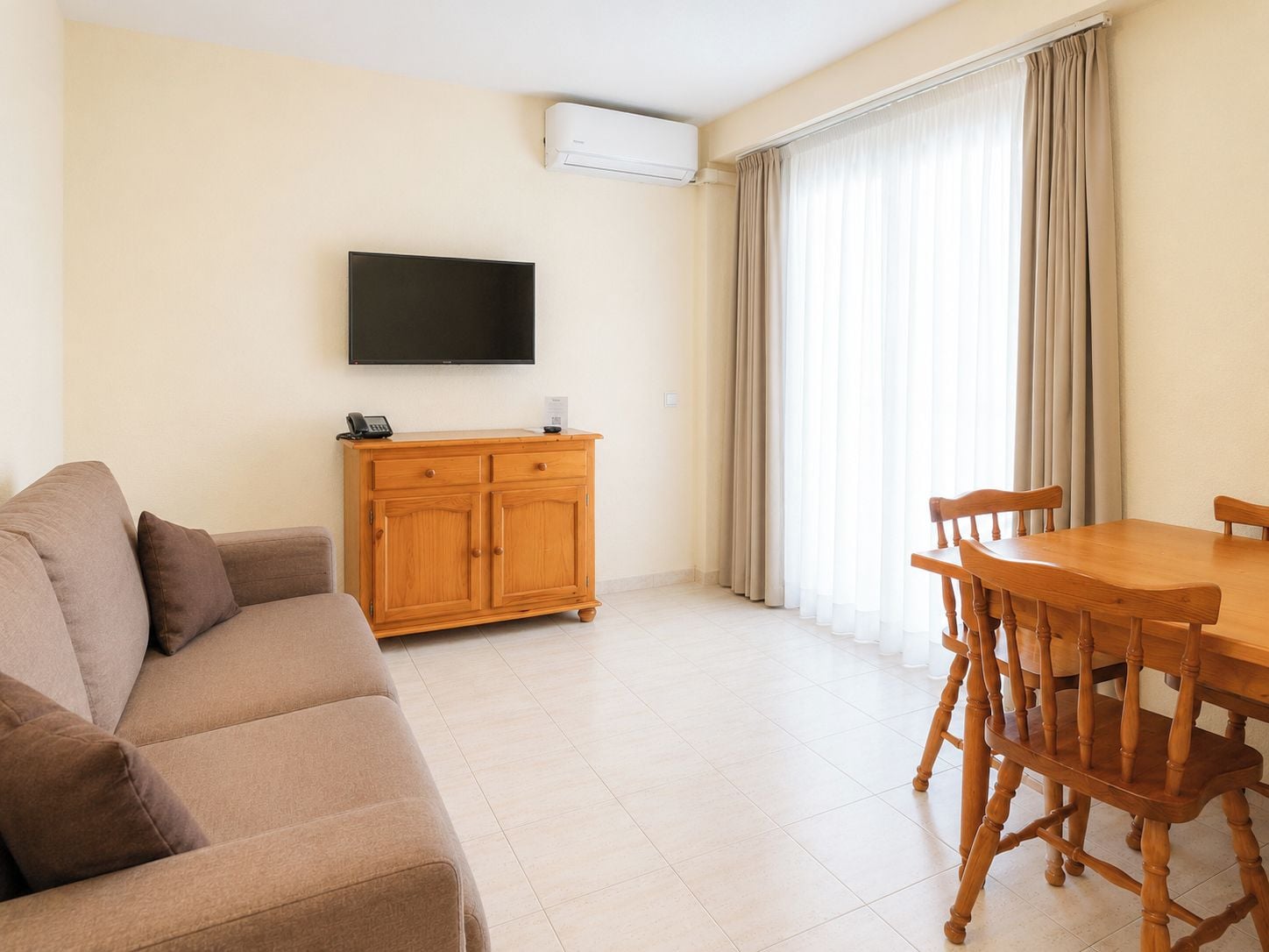 A living room with a sofa, a wooden cabinet, a flat-screen TV, a dining table with chairs, and a window with curtains.