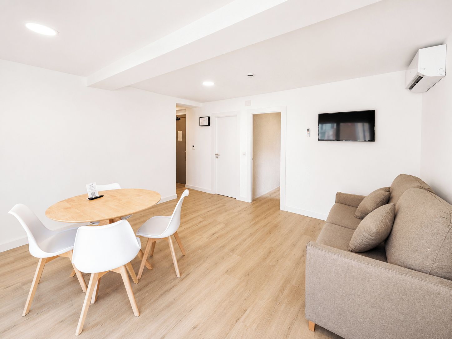 A modern living room with a round wooden table, white chairs, a beige sofa, and a mounted TV.