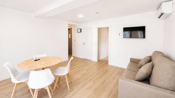 A modern living room with a round wooden table, white chairs, a beige sofa, and a mounted TV.