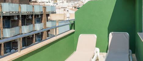 A rooftop terrace with two lounge chairs, a green wall, and a view of buildings and a mountain in the distance.
