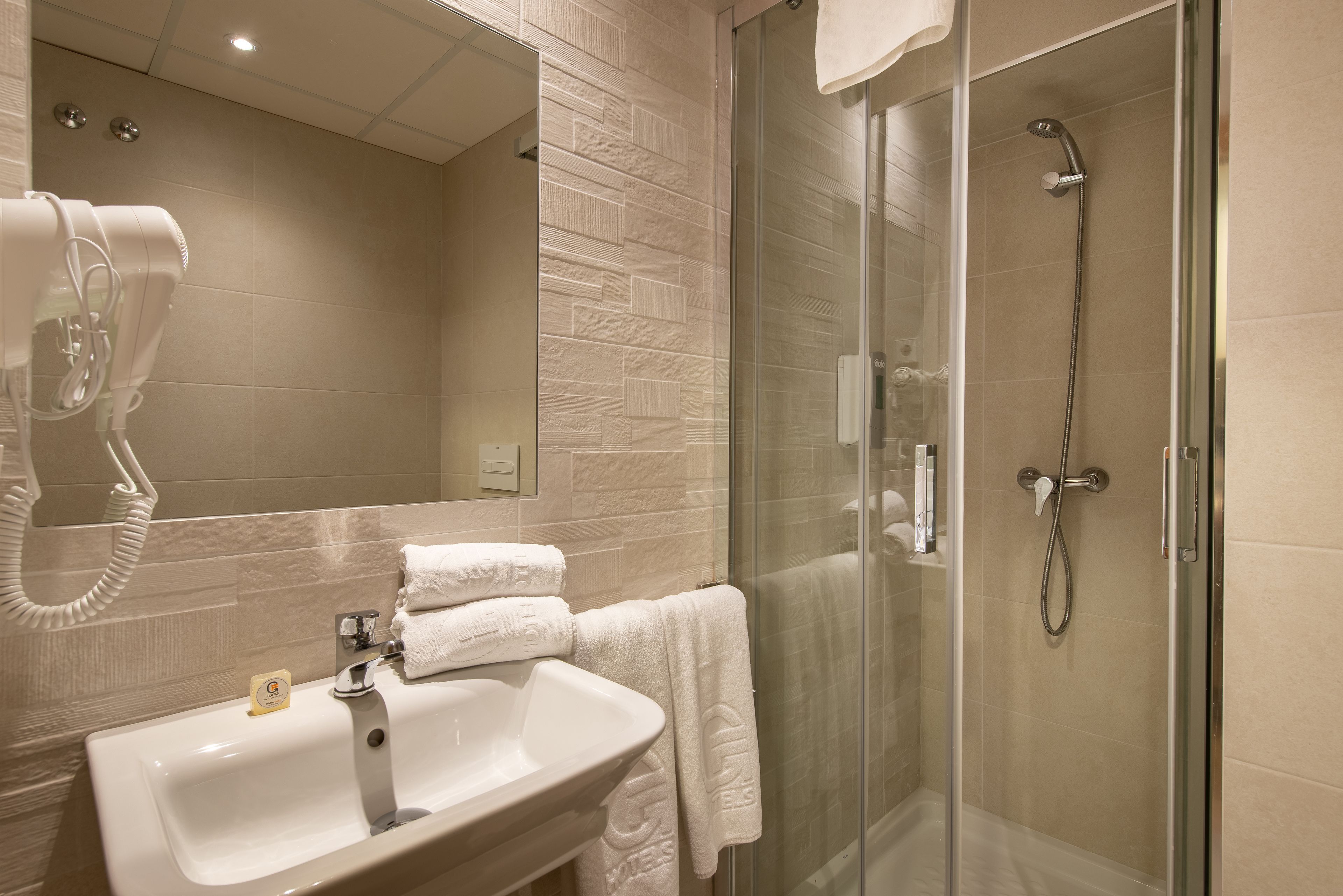 Ein modernes Badezimmer mit einer ein Glasfach trennenden Duschkabine, einem weißen Waschbecken mit Seifenspender und einem Fön auf einem Wandhalterung.