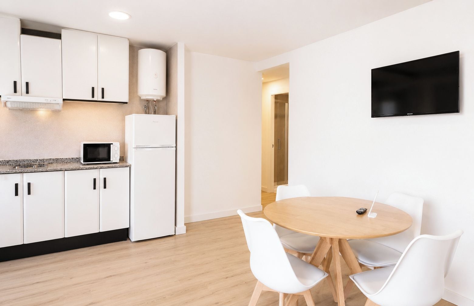 A modern kitchen with white cabinets, a microwave, a refrigerator, and a water heater. There is a round wooden table with white chairs and a flat-screen TV mounted on the wall.
