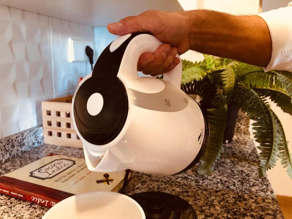 Una mano sostiene una cafetera blanca y negra sobre una encimera de cocina, con un libro de recetas y una planta al fondo.