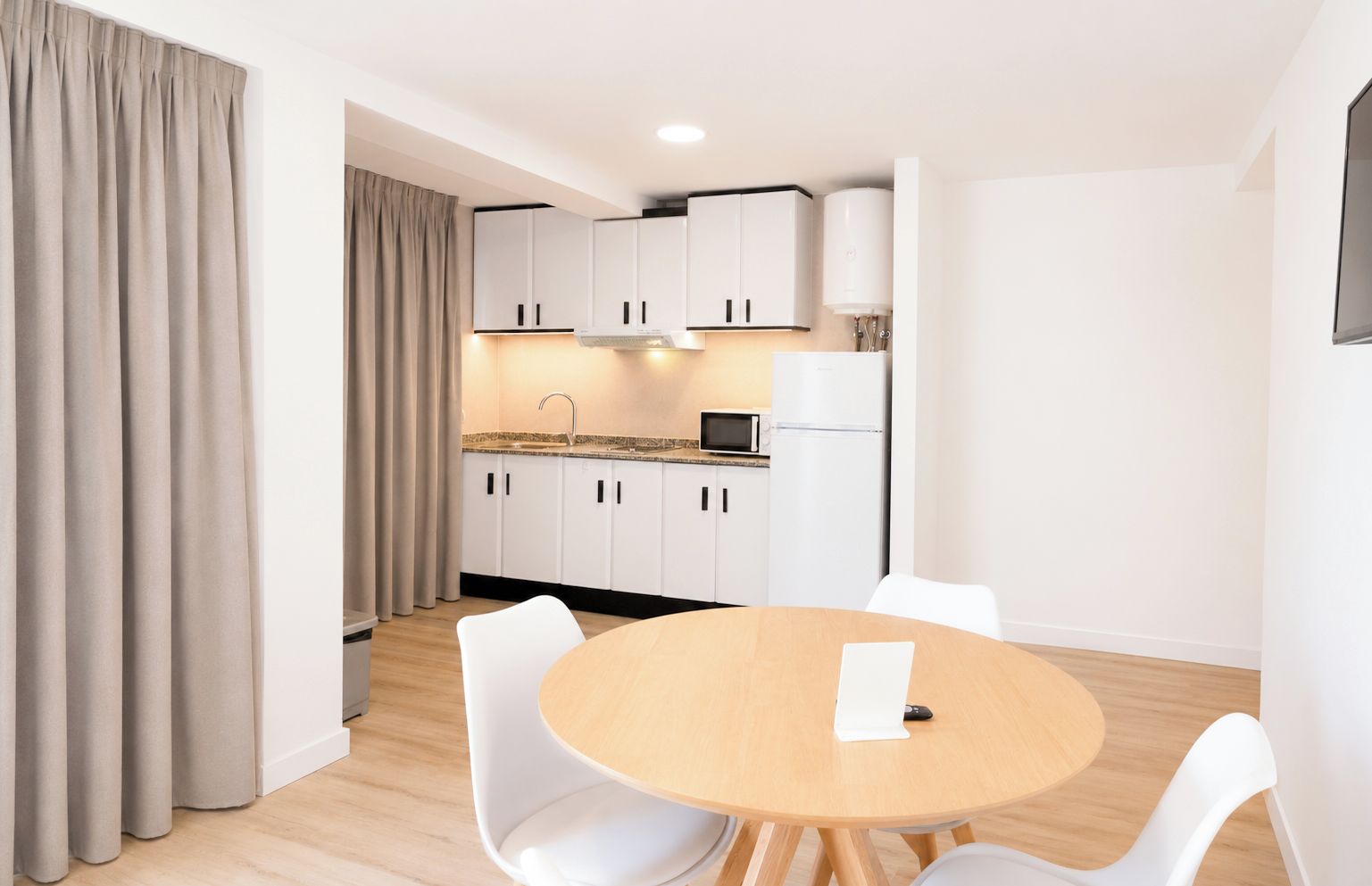 A modern kitchen with white cabinets, a microwave, and a refrigerator. There is a round wooden table with white chairs in the foreground.