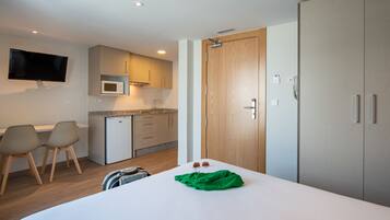Habitación de hotel con cama, escritorio con sillas, kitchenette y televisor montado en la pared.