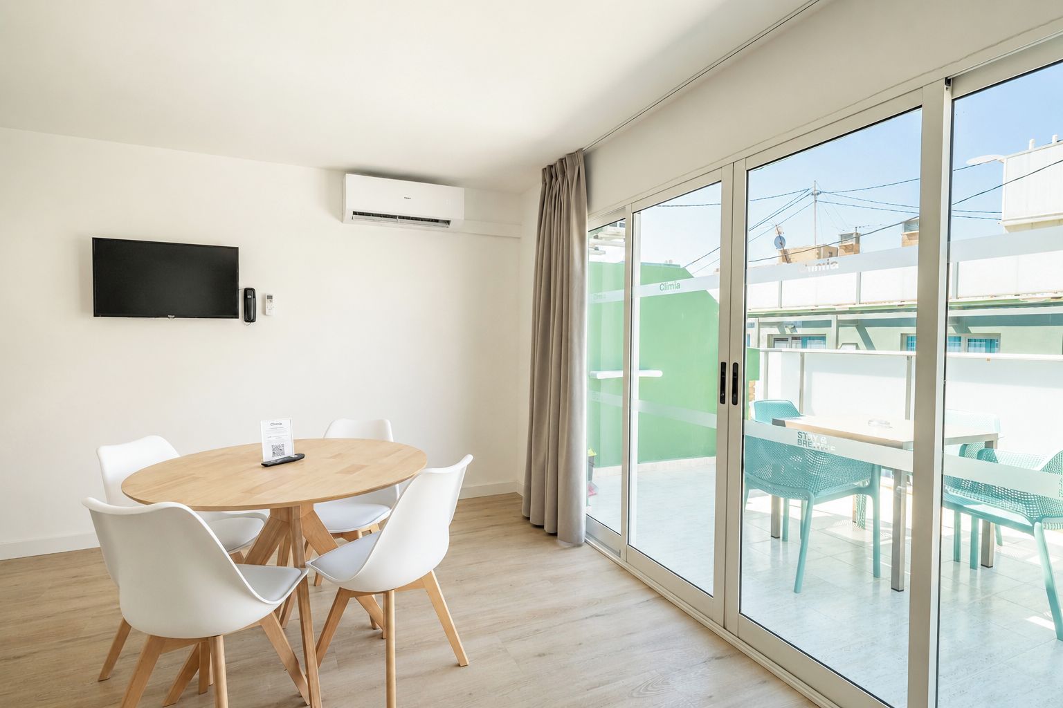 A modern room with a round wooden table, white chairs, a flat-screen TV, and a large sliding glass door leading to an outdoor area with green chairs.
