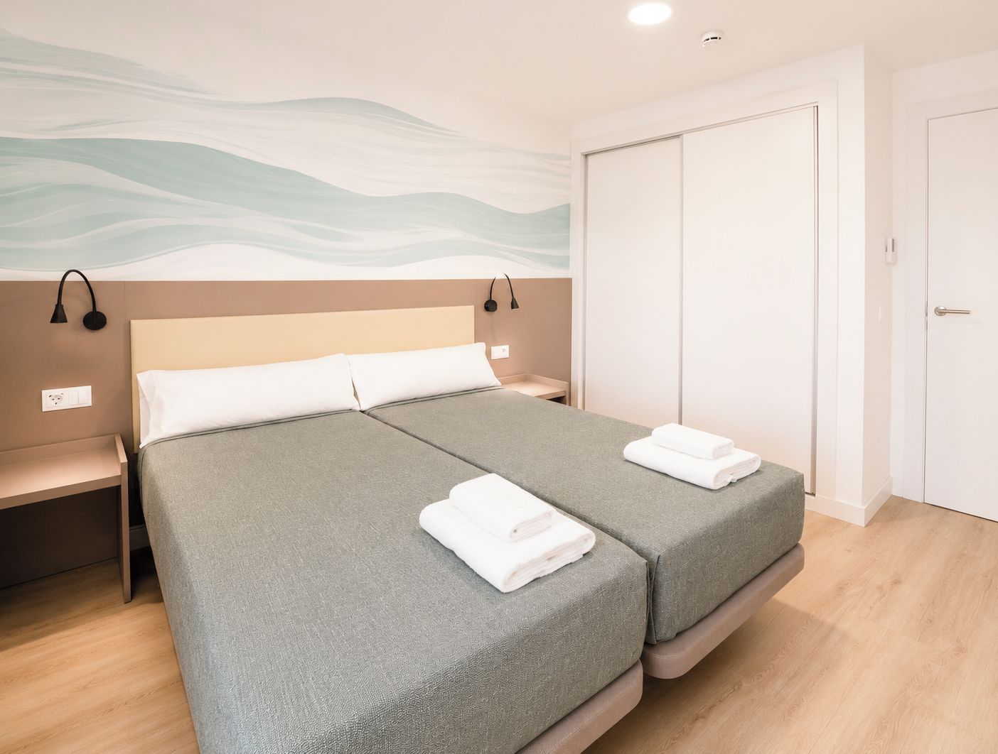 A double bed with grey bedding, a bedside table, a wall-mounted lamp, and a wardrobe.