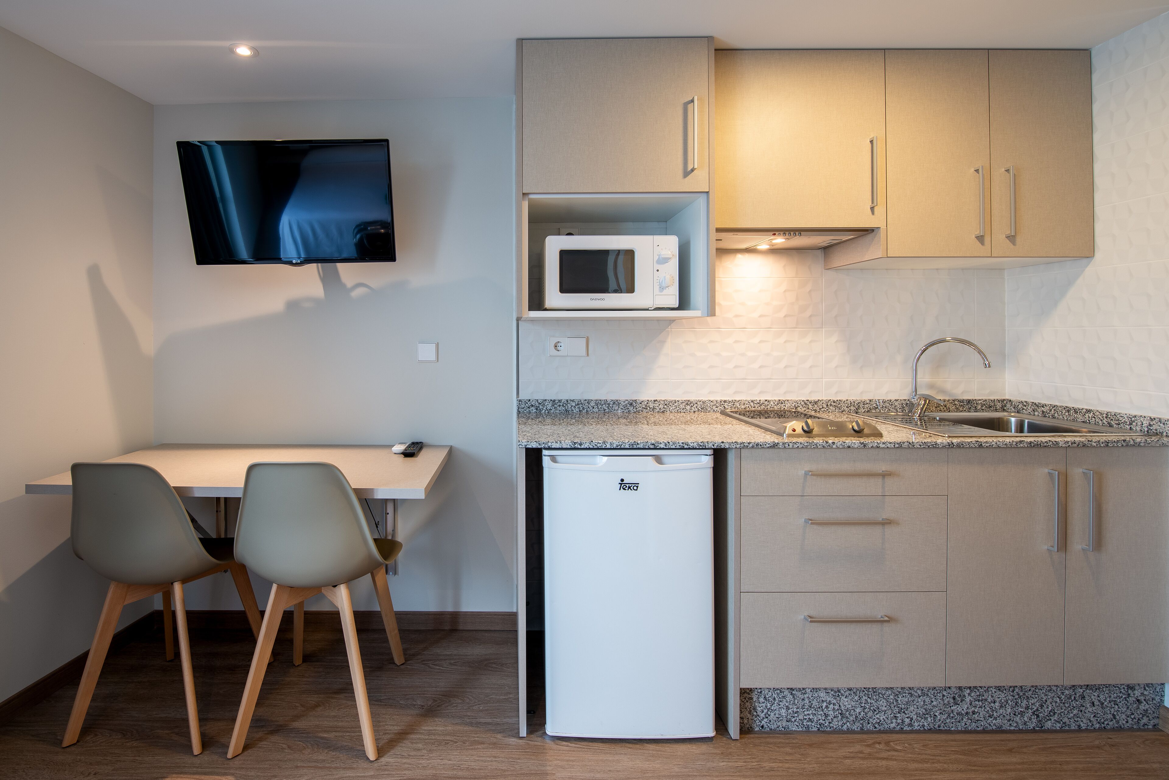 A compact kitchen with a microwave, sink, and a small refrigerator. There is a dining area with two chairs and a table. A flat-screen TV is mounted on the wall.