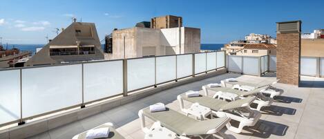 A rooftop terrace with sun loungers, a glass balustrade, and a view of the sea and buildings.