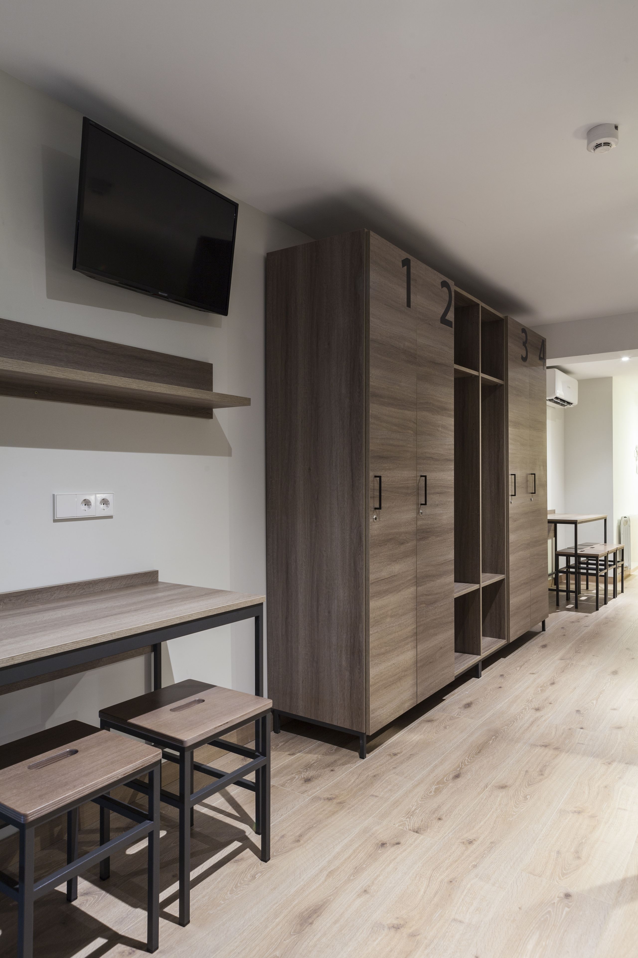 Una habitación moderna con un armario de madera, un televisor de pantalla plana en la pared y una pequeña mesa con dos taburetes.