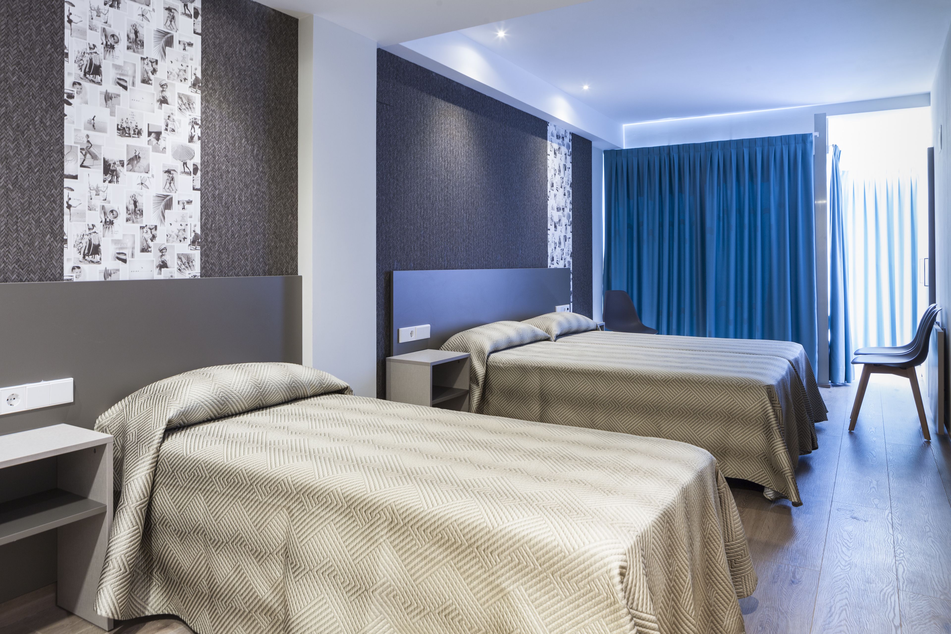 A hotel room with two beds, a desk, a chair, and a wall with a patterned wallpaper.