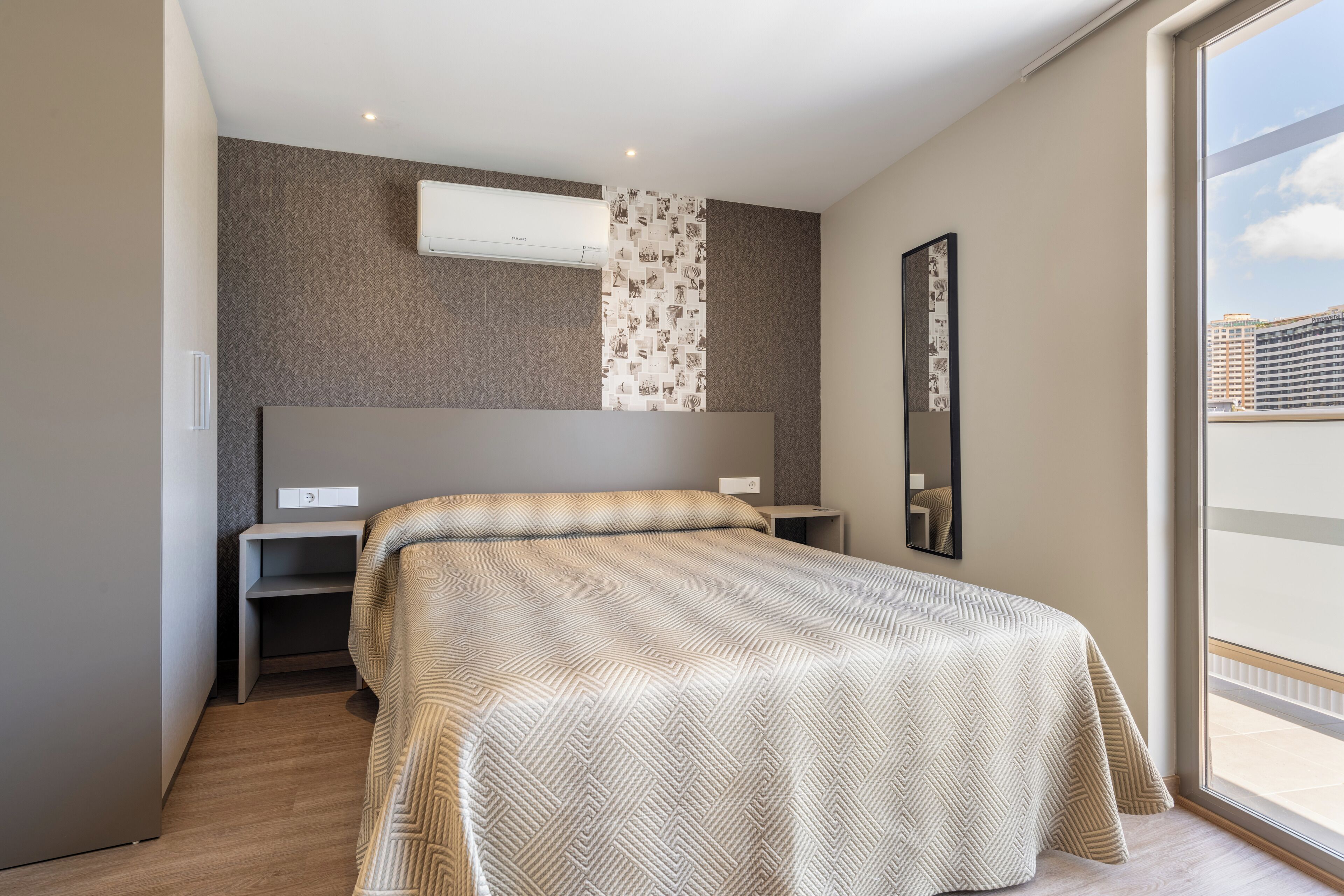 A bedroom with a large bed, a desk, a chair, a wardrobe, and a window with a view of buildings.