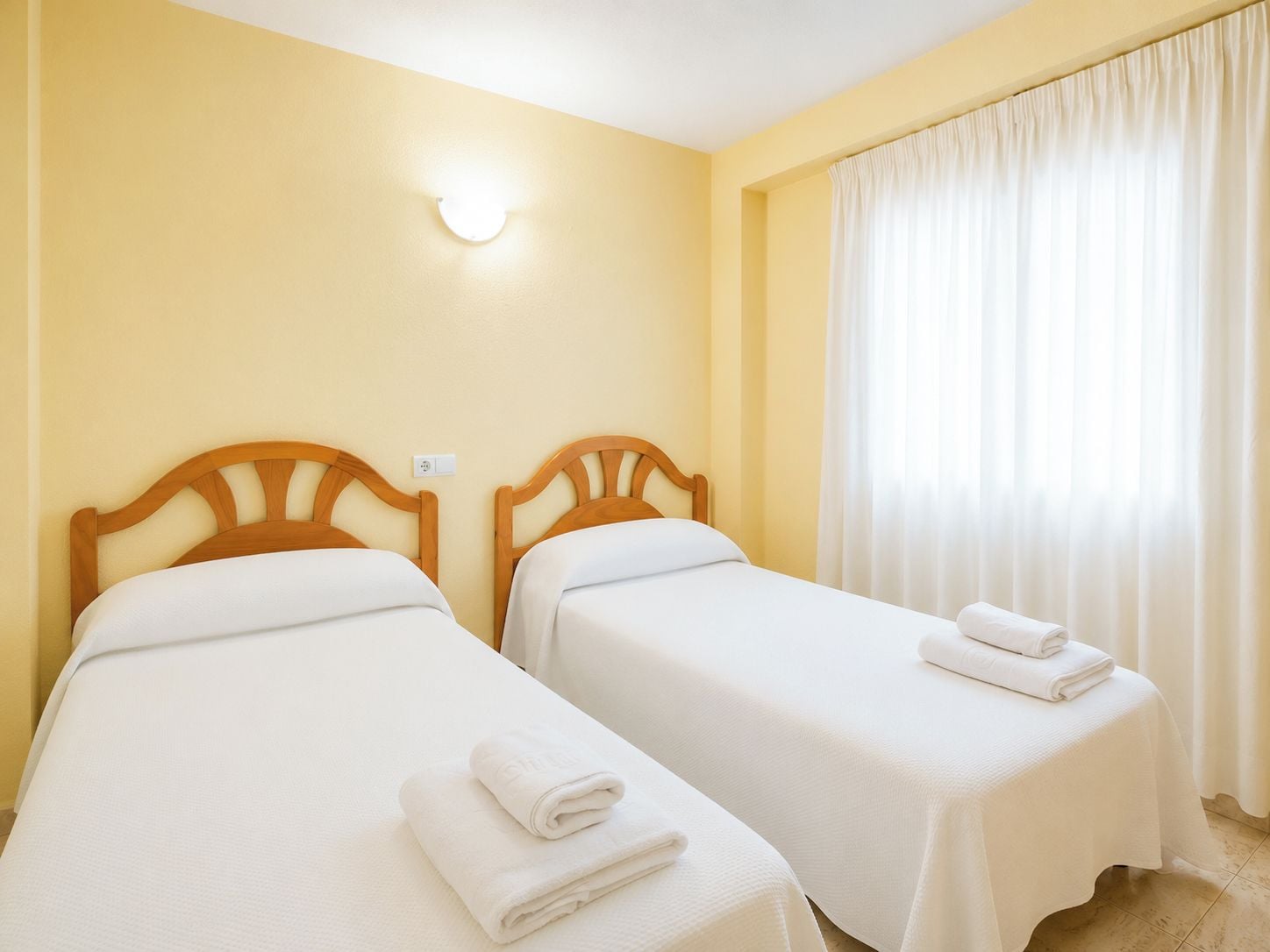 Una habitación de hotel con dos camas individuales, una mesita de noche con una toalla enrollada y una ventana con cortinas.