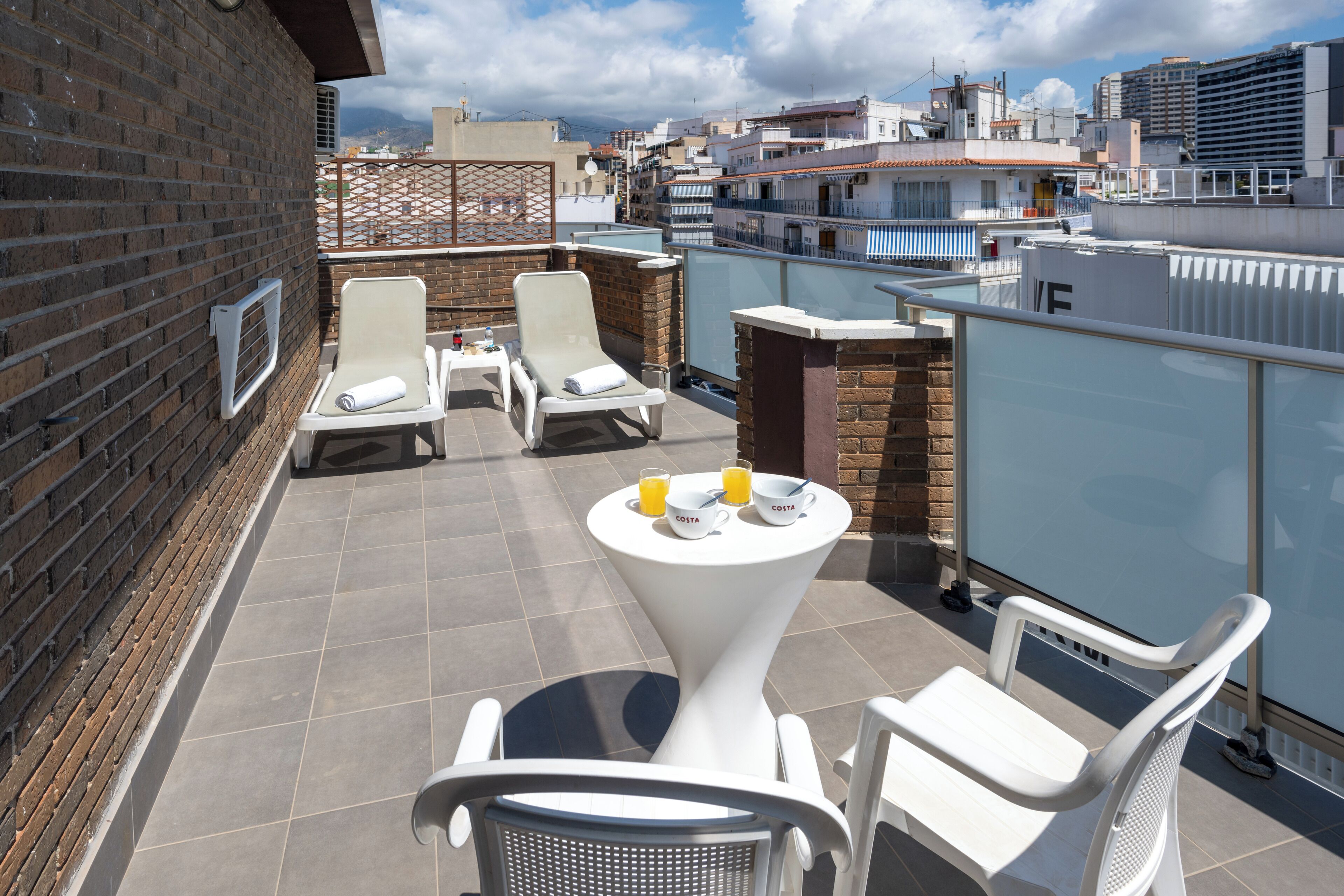 A rooftop terrace with white lounge chairs, a table with a glass of orange juice, and a view of the cityscape.