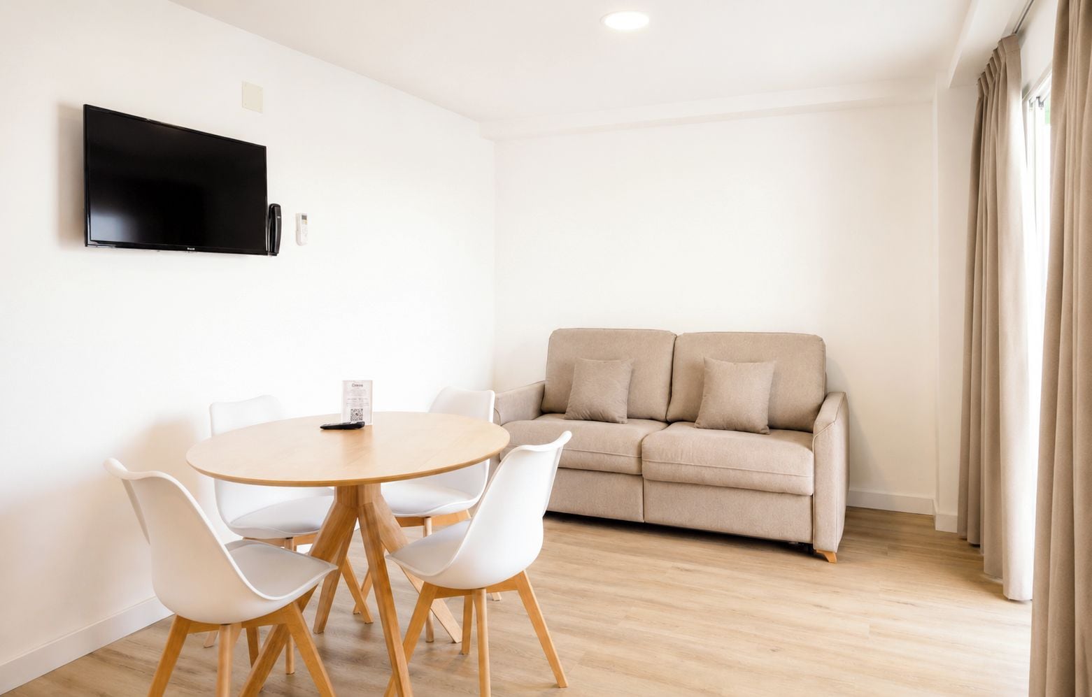 A modern living room with a beige sofa, a round wooden table with chairs, a flat-screen TV mounted on the wall, and a window with curtains.