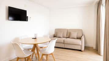 A modern living room with a beige sofa, a round wooden table with chairs, a flat-screen TV mounted on the wall, and a window with curtains.