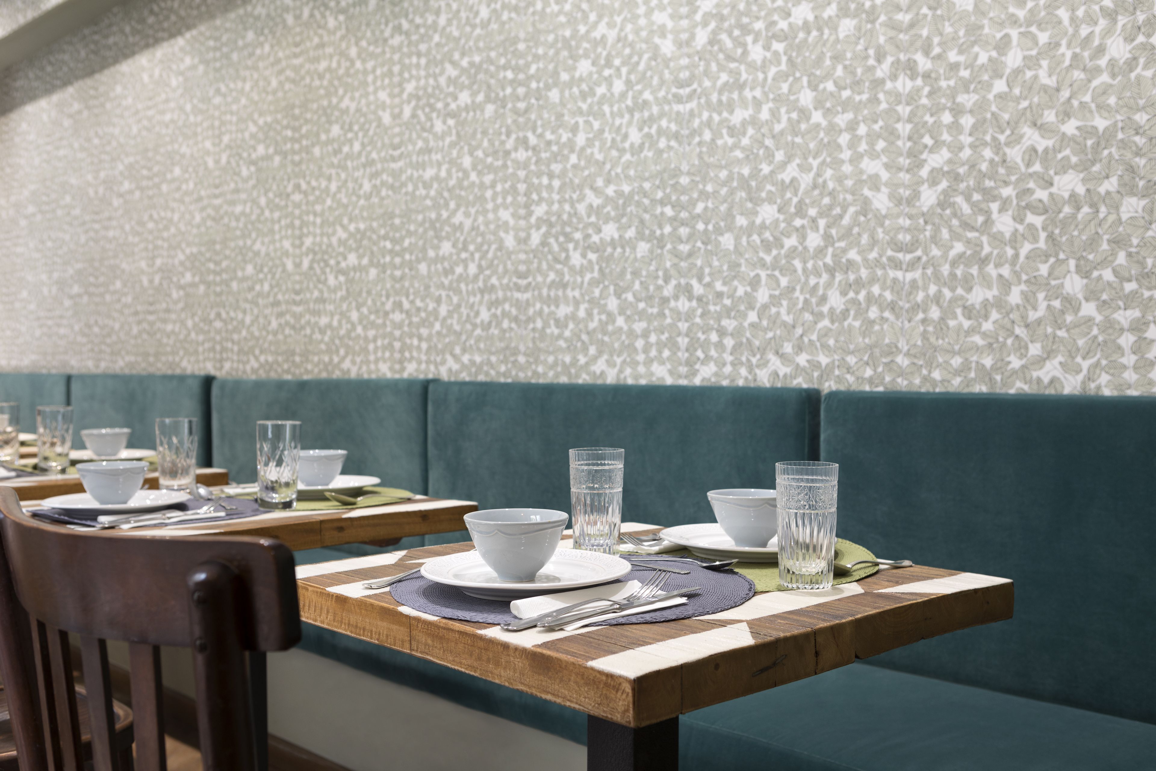 Un espace repas avec des tables préparées pour deux personnes, agrémenté d’un mur à motifs et d’un banc recouvert d’un tissu turquoise.