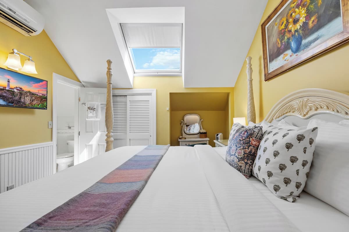 A bedroom with a large bed, a skylight, a painting, and a view of the bathroom.