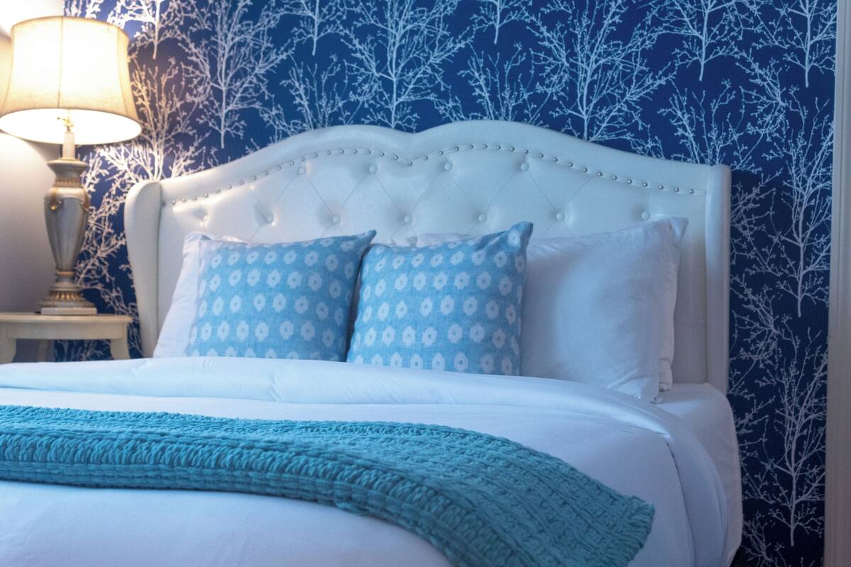 A bedroom with a white bed, two patterned pillows, a teal throw blanket, and a nightstand with a lamp.