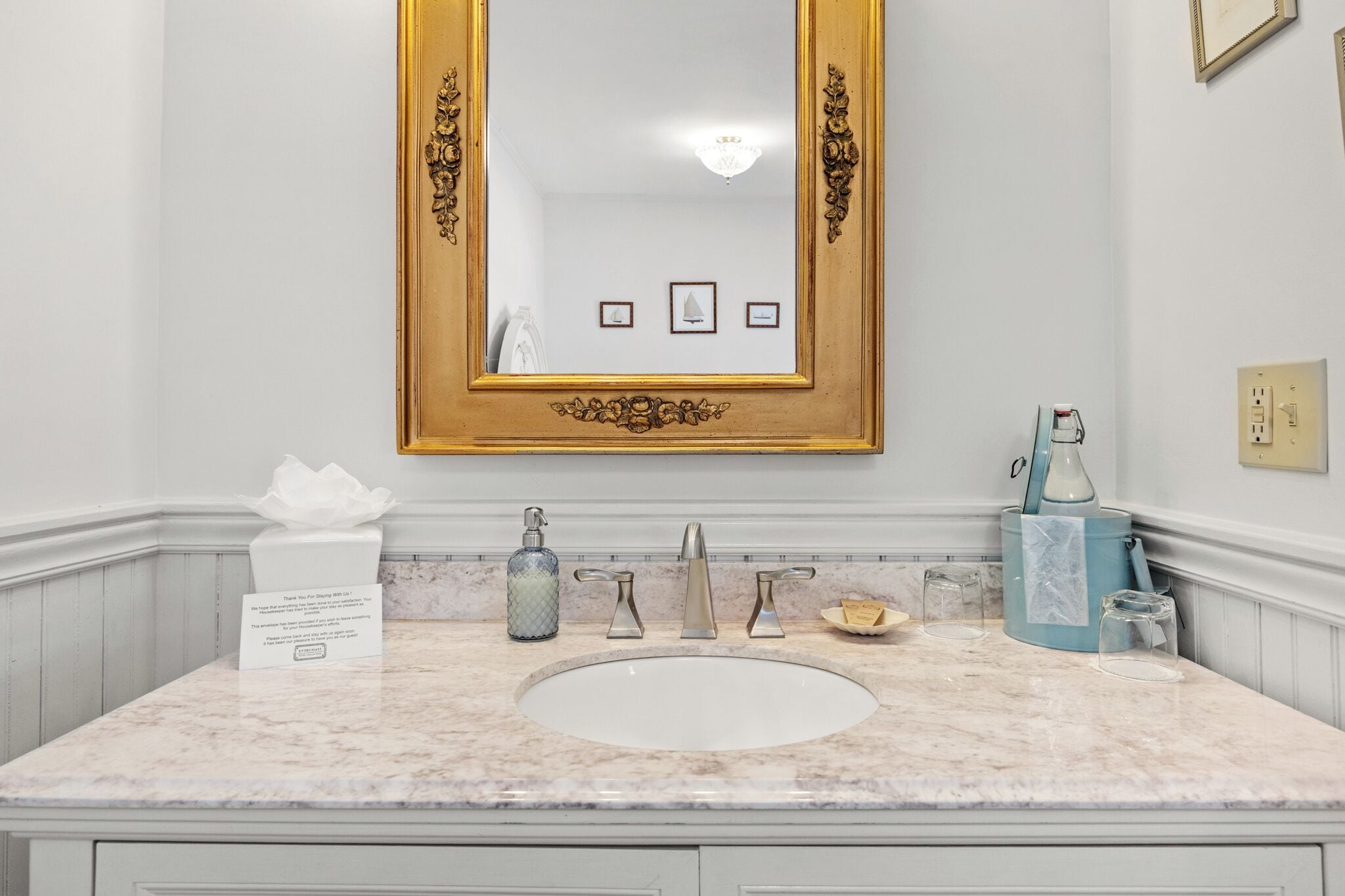 A bathroom with a marble sink, gold-framed mirror, and a towel dispenser.