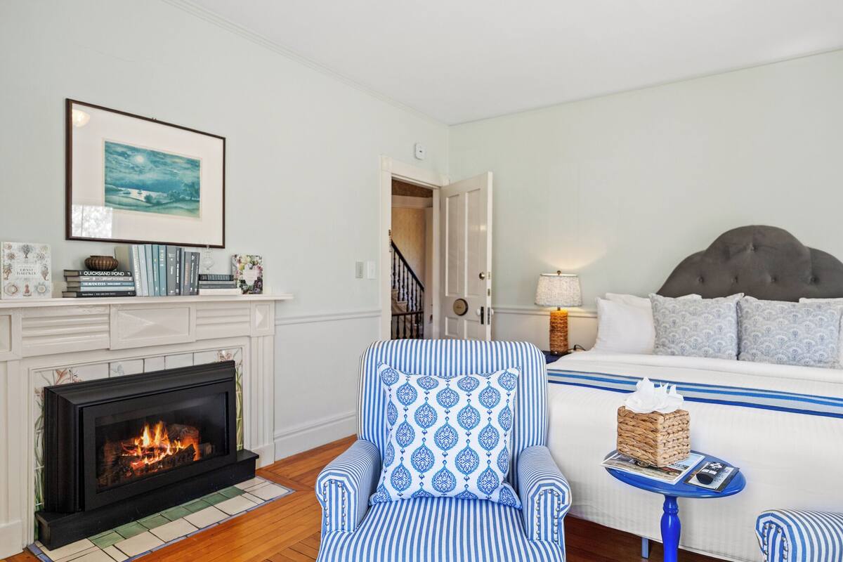 A hotel room with a fireplace, a bed with a headboard, a striped armchair, and a small table with a lamp.
