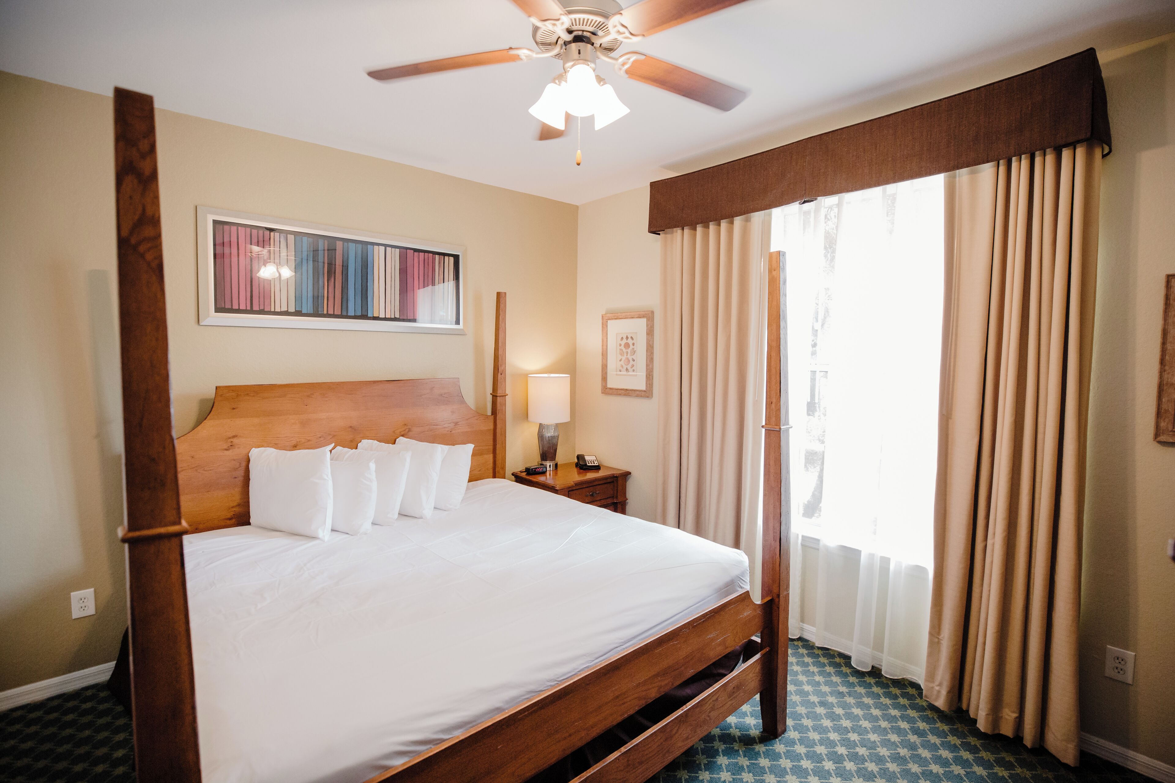 Une chambre d’hôtel avec un lit à baldaquin, un ventilateur de plafond, une grande fenêtre avec des rideaux et une œuvre d’art accrochée au mur.